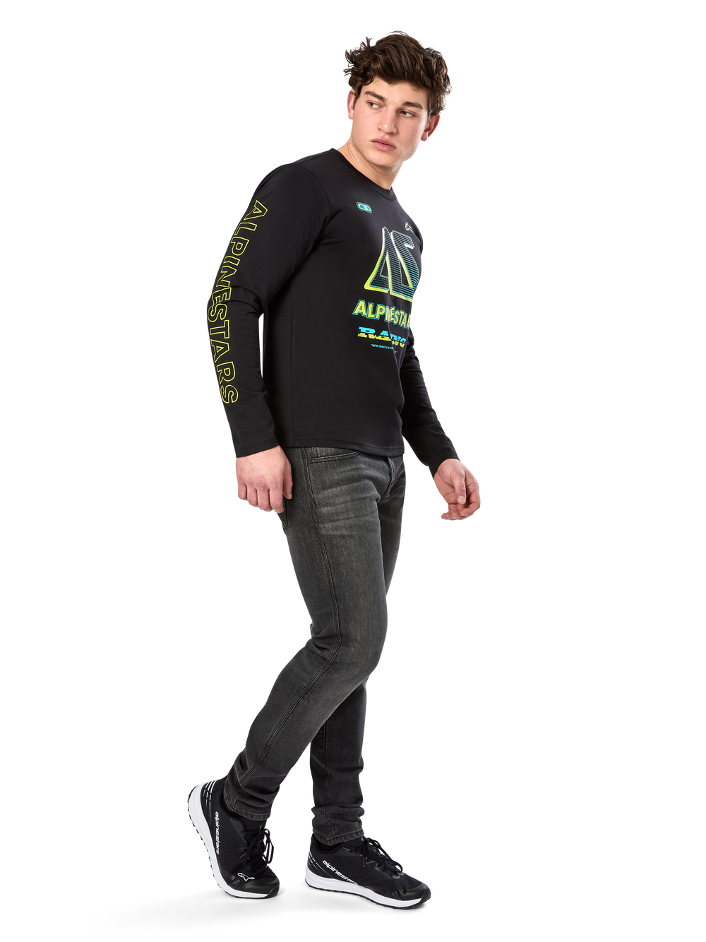 Man wearing Alpinestars Ayess CSF Long Sleeve Tee, Black, featuring yellow sleeve lettering and vibrant lime and cyan chest graphics, paired with dark grey jeans and black sneakers, casual motorsport-inspired streetwear.