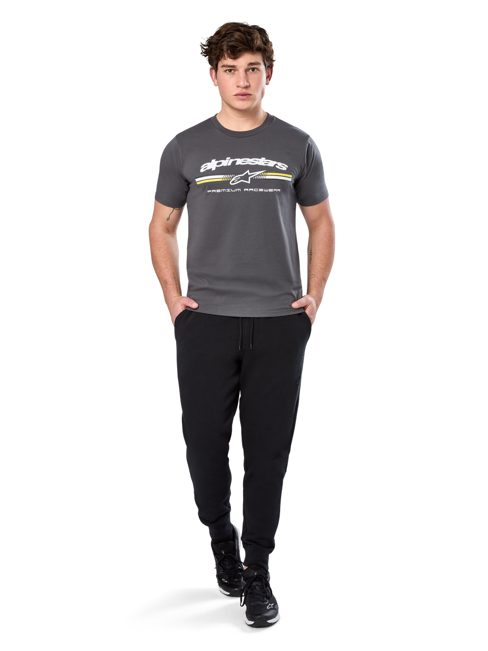 Man wearing Alpinestars Prevail Csf Tee, Short Sleeve T-Shirt, Charcoal, featuring white Alpinestars logo and tri-color stripe with yellow accents, paired with black joggers and sneakers, casual motorsport streetwear