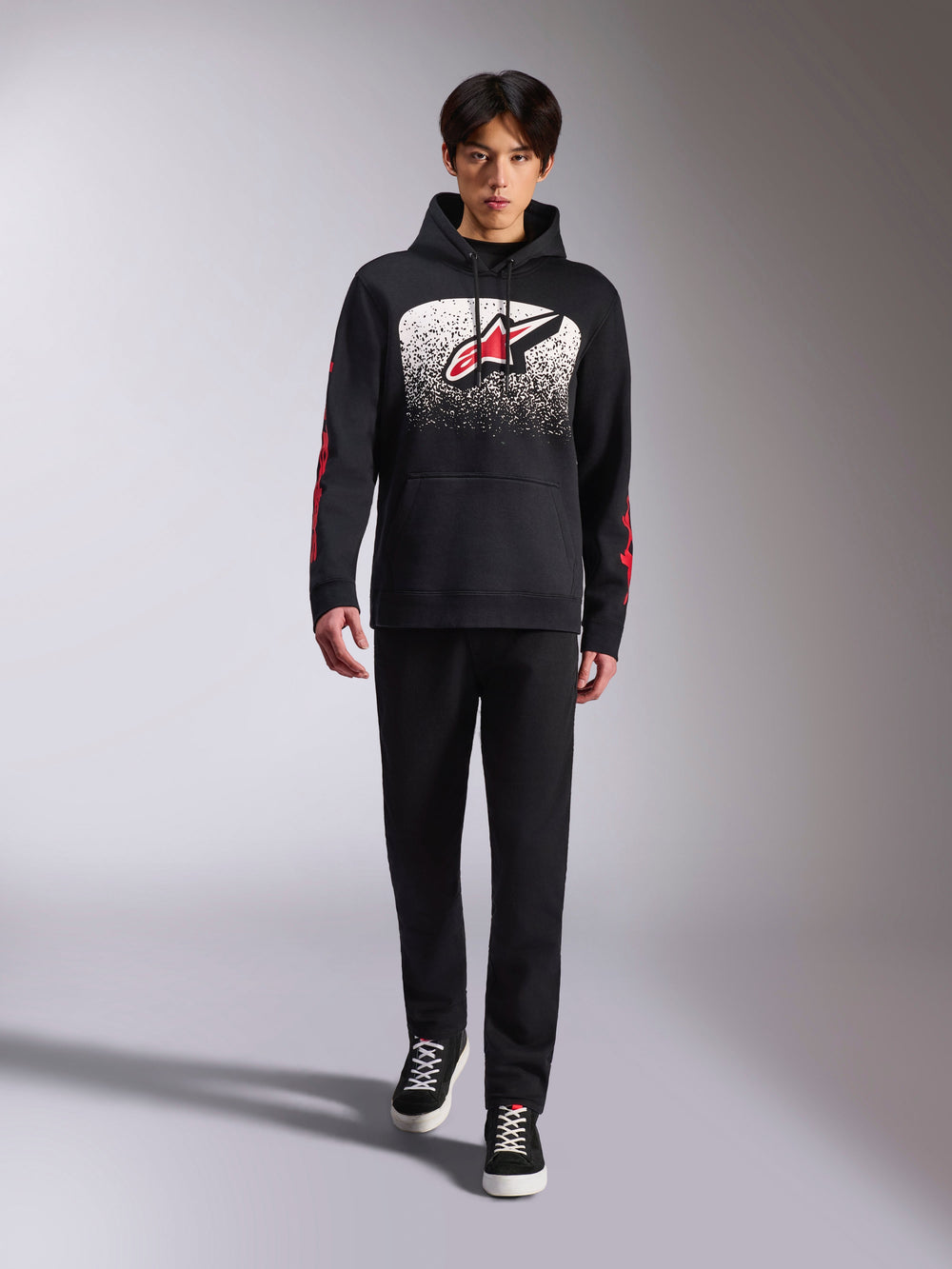 Alpinestars Obsessed Hoodie, black pullover hoodie, large white and red Astars logo with MX gradation, red sleeve wordmarks, worn by model in a full black casual outfit, motorsport-inspired lifestyle apparel