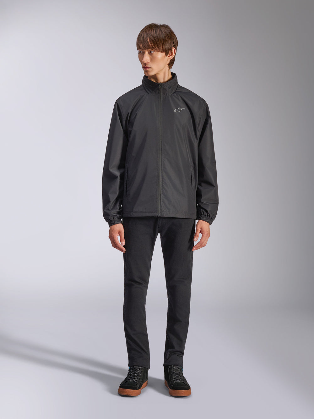 Alpinestars Overhaul Racer Jacket, Black Men's Rain Shell, full-length view on a model paired with black pants and casual sneakers, featuring a small white logo on the chest, water-resistant fabric, and casual motorsport-inspired design