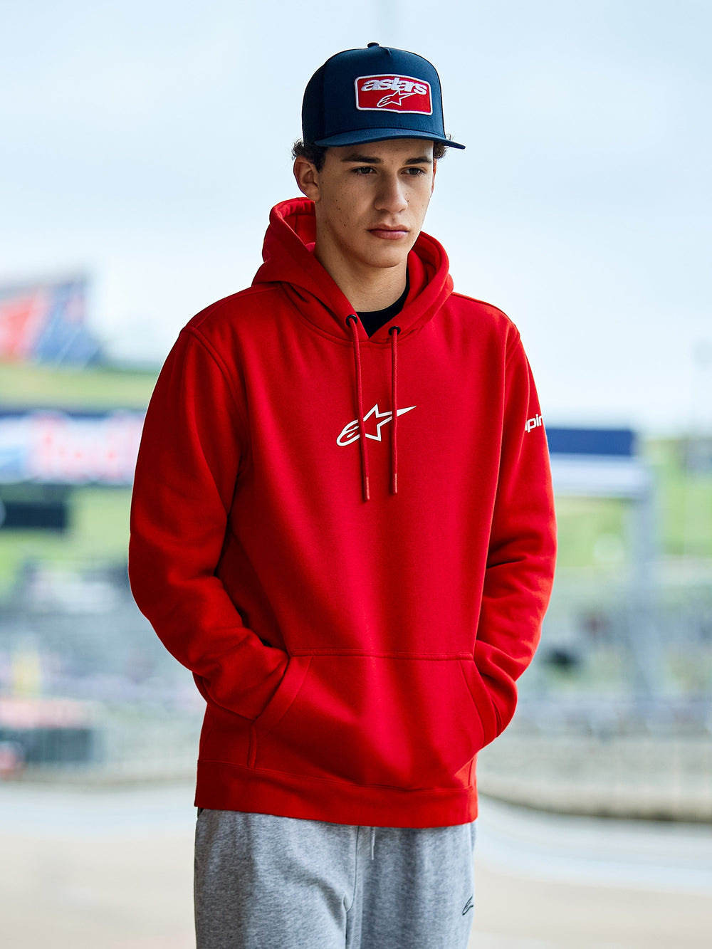 Alpinestars Frontal Hoodie, Red pullover fleece hoodie, featuring a white Astars logo on the chest, kangaroo pocket, and drawstring hood, worn by a man in a blue cap, casual motorsport streetwear