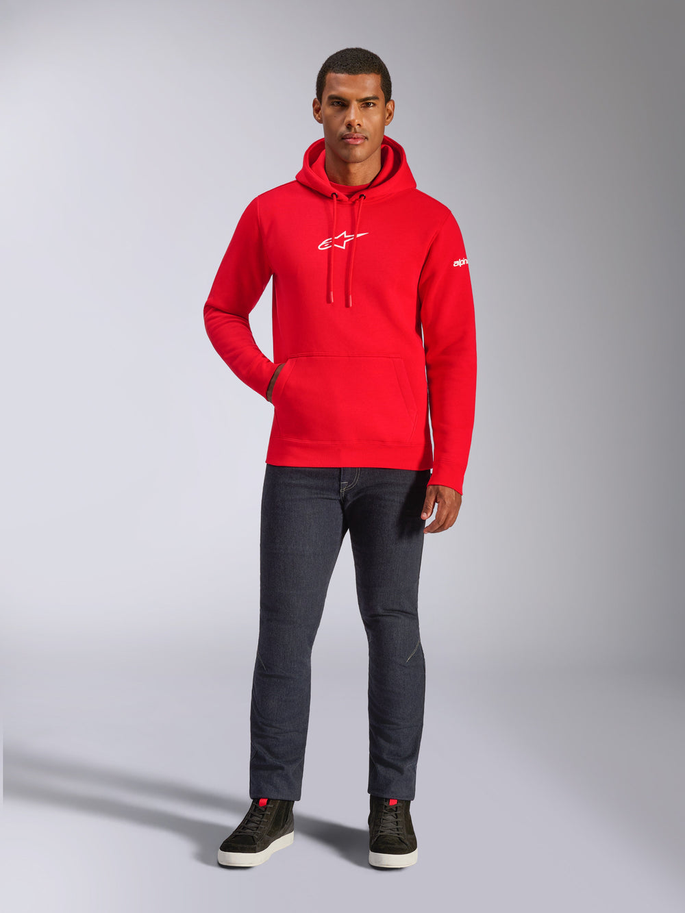 Male model wearing Alpinestars Frontal Hoodie in Red, pullover fleece hoodie with white Astars logo on chest, kangaroo pocket, drawstring hood, paired with dark grey jeans and black high-top sneakers, casual motorsport streetwear