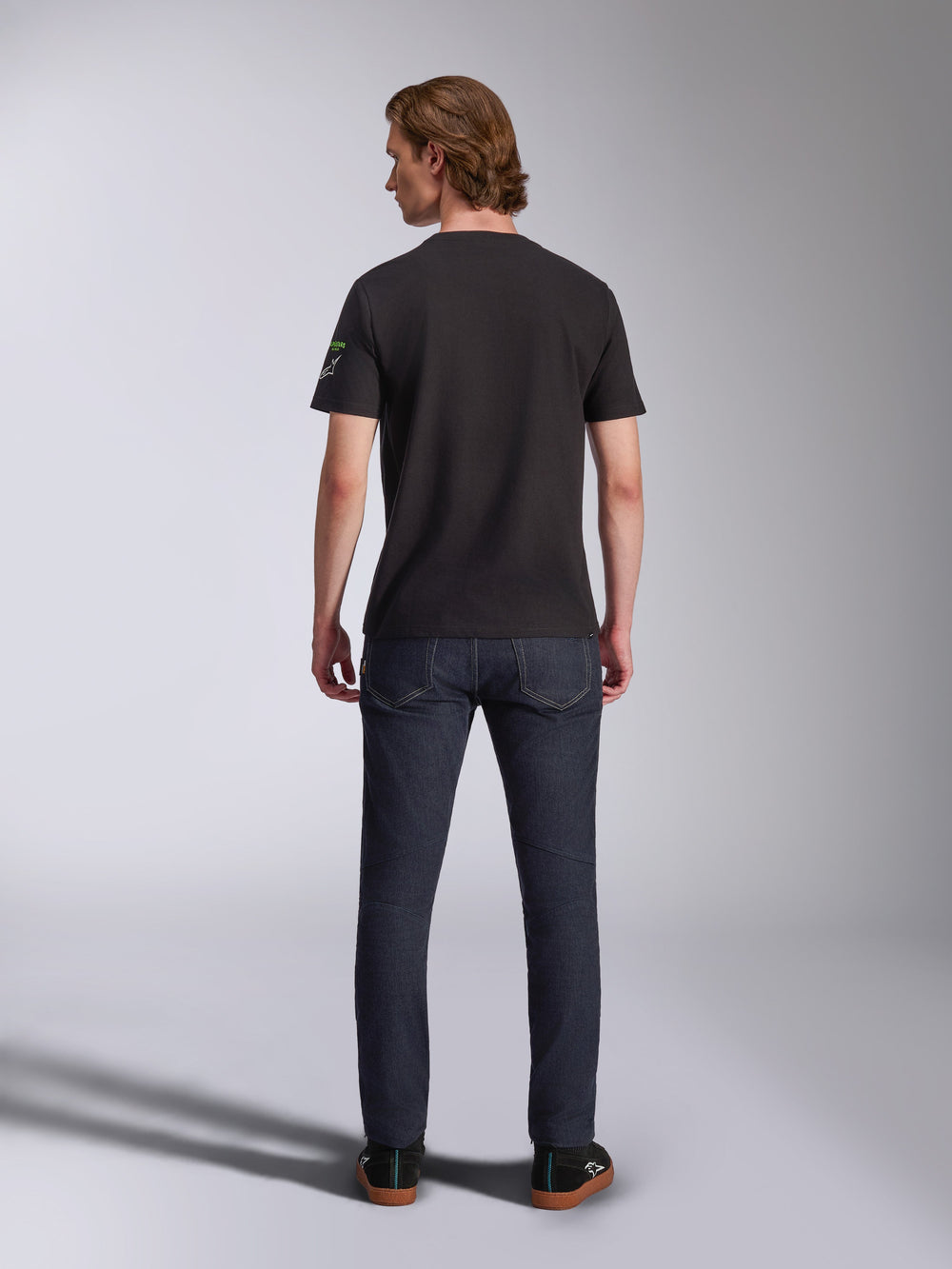 Alpinestars Entitled CSF Tee, Short Sleeve T-Shirt, Black, back view of a male model wearing the shirt with dark blue denim jeans, featuring a small white and green logo on the left sleeve, casual motorsport-inspired streetwear