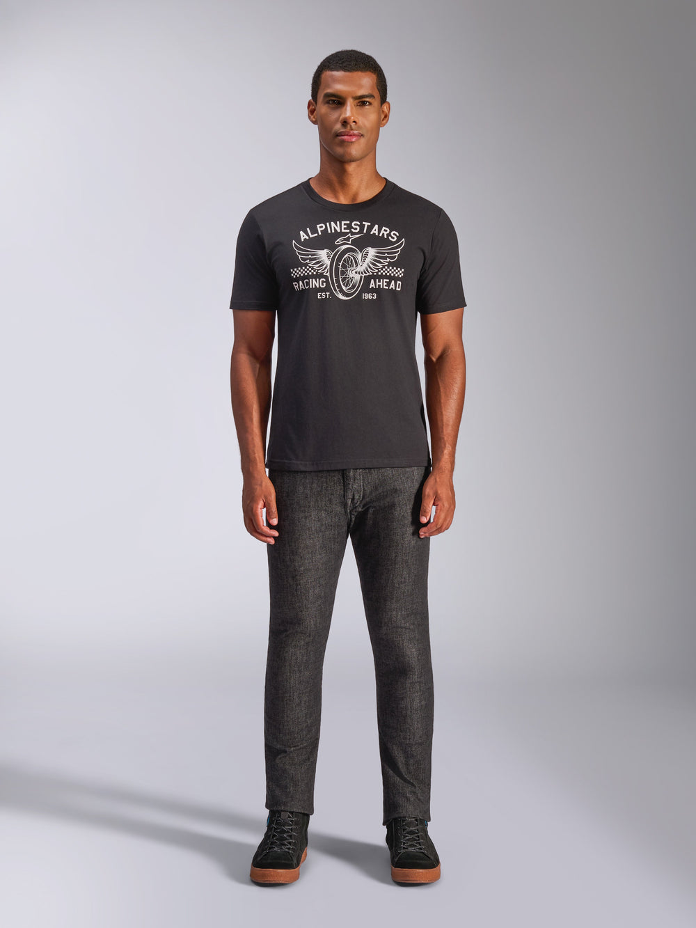 Alpinestars Heritage Patch CSF Tee, Short Sleeve T-Shirt, Black, featuring a white winged wheel graphic with 'Racing Ahead' text, worn by a model with dark grey pants and black sneakers, Casual Motorsport Streetwear