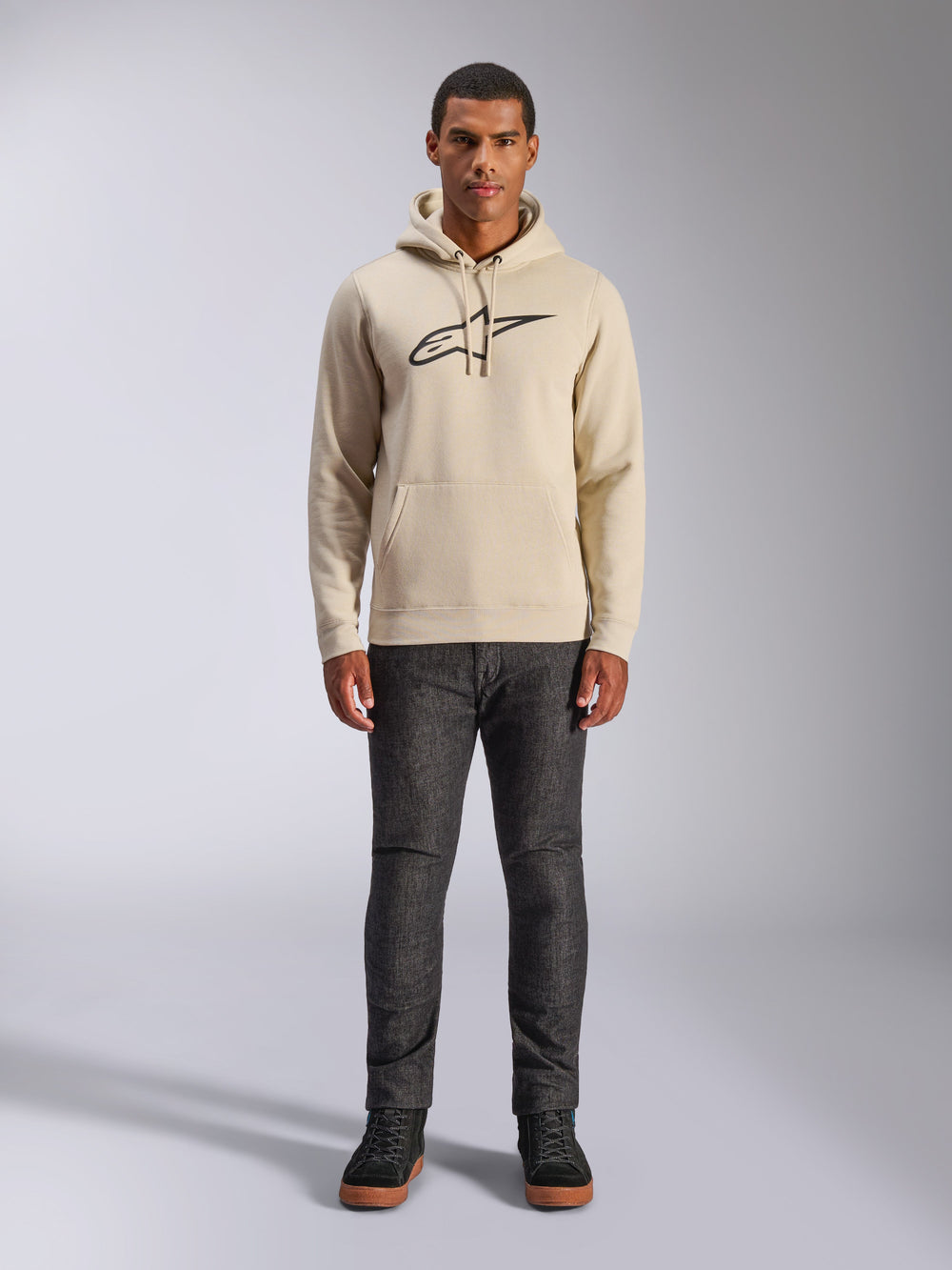 Alpinestars Ageless V3 Hoodie, casual sportswear, Off White with black embroidered logo, worn by a man with dark grey pants and black sneakers, featuring a drawstring hood and kangaroo pocket, modern motorsport-inspired design