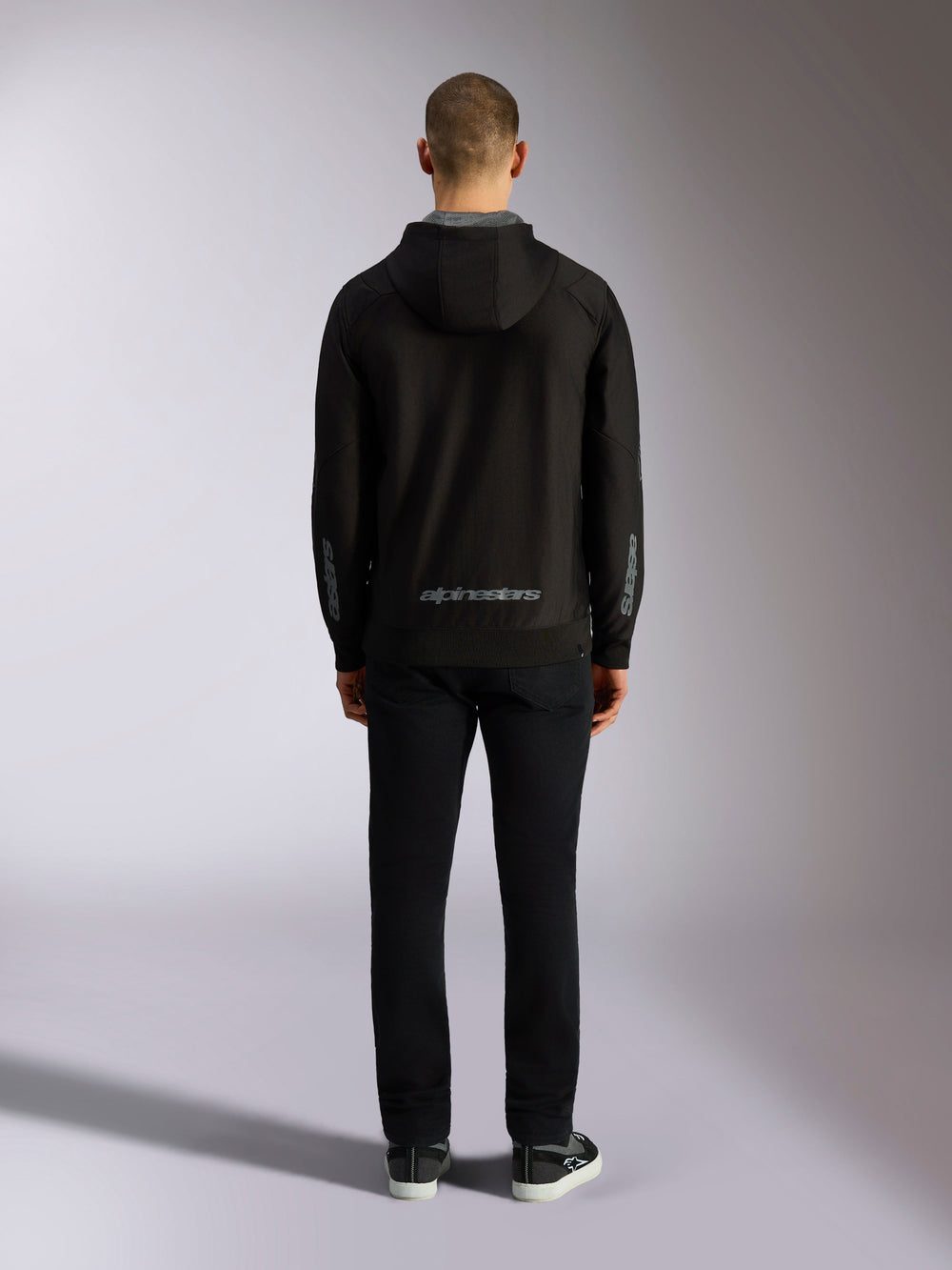 Back view of Alpinestars Sessions V3 Hoodie in Black Grey, featuring a grey Alpinestars wordmark on the lower back and astars logos on the forearms, worn by a model with black pants, casual motorsport-inspired streetwear