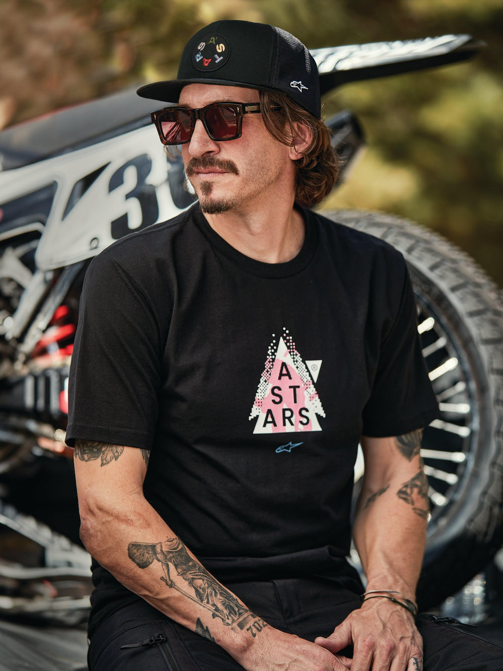 Alpinestars Gambit Trucker Hat in black with circular logo patch, worn with Alpinestars Soloist SS Tee in black, lifestyle shot featuring a man with sunglasses and tattoos sitting in front of a motocross bike.