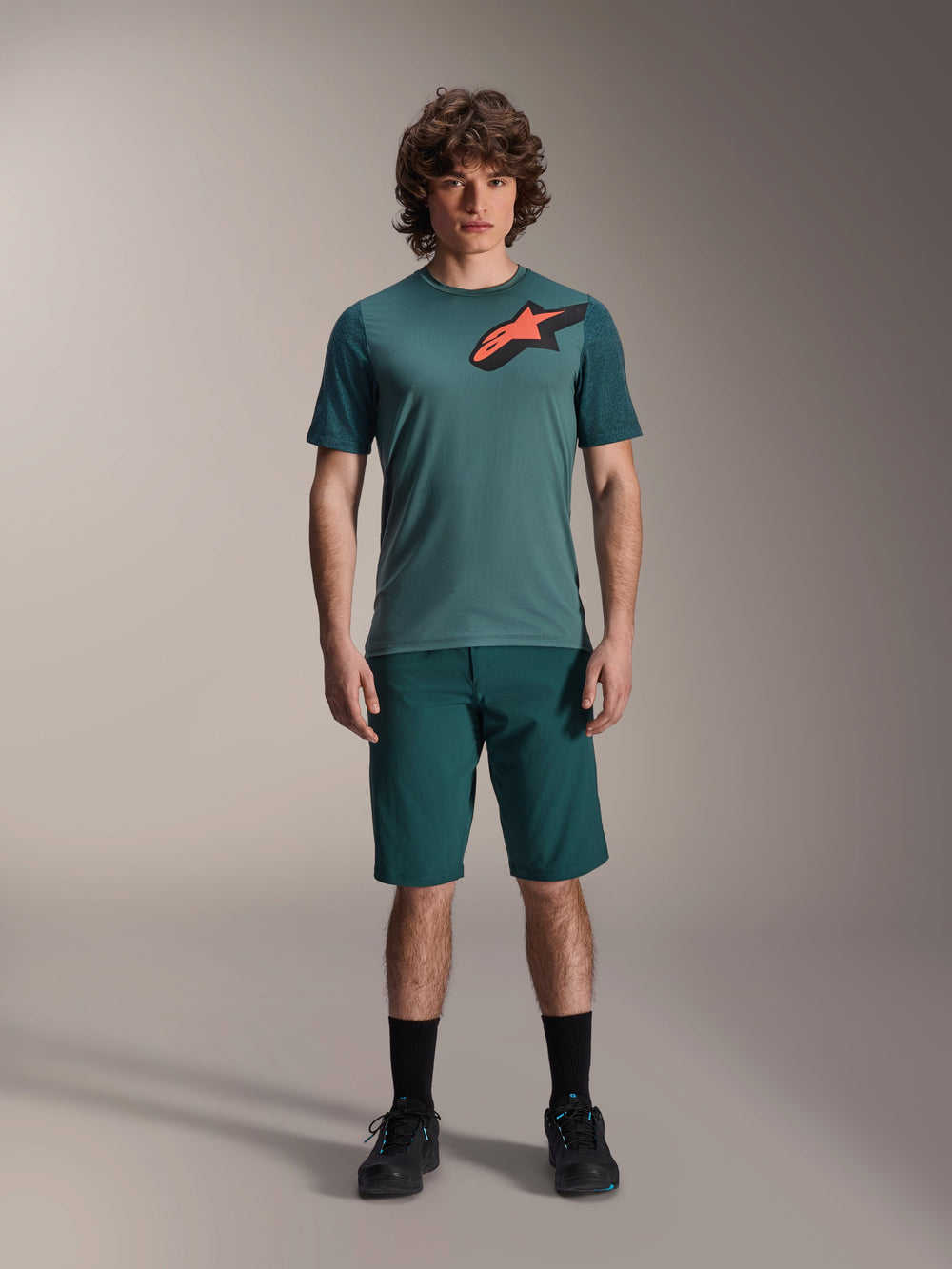 Alpinestars A-Dura Shorts in Dark Pine, men's mountain bike shorts, shown on a model with a matching teal short-sleeve jersey featuring a red and black star logo, black socks and cycling shoes, casual sporty mountain biking apparel