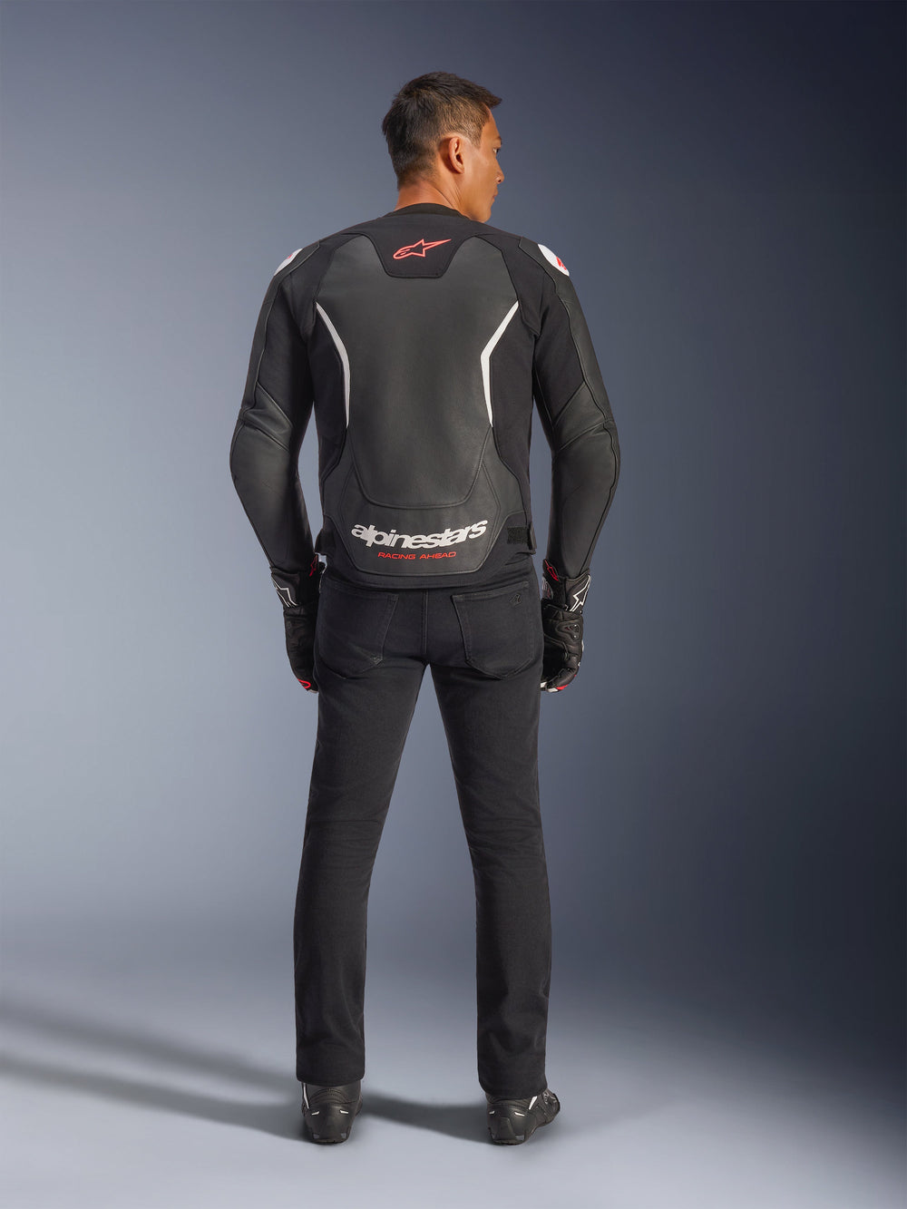 Alpinestars GP Force V2 Leather Jacket, Black and White, rear view, worn by a rider with black pants and gloves, featuring large white branding on the lower back and red accents, premium bovine leather construction for motorcycle road gear