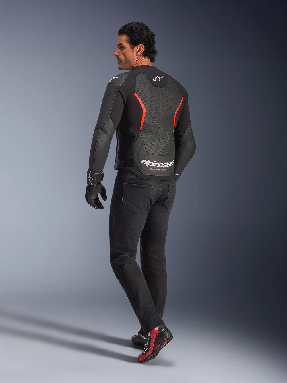 Male model from the back wearing the Alpinestars GP Force V2 Air Leather Jacket in Black Red Fluo, featuring extensive back perforations and bold white branding, paired with black riding jeans, gloves, and black and red riding boots in a studio setting.