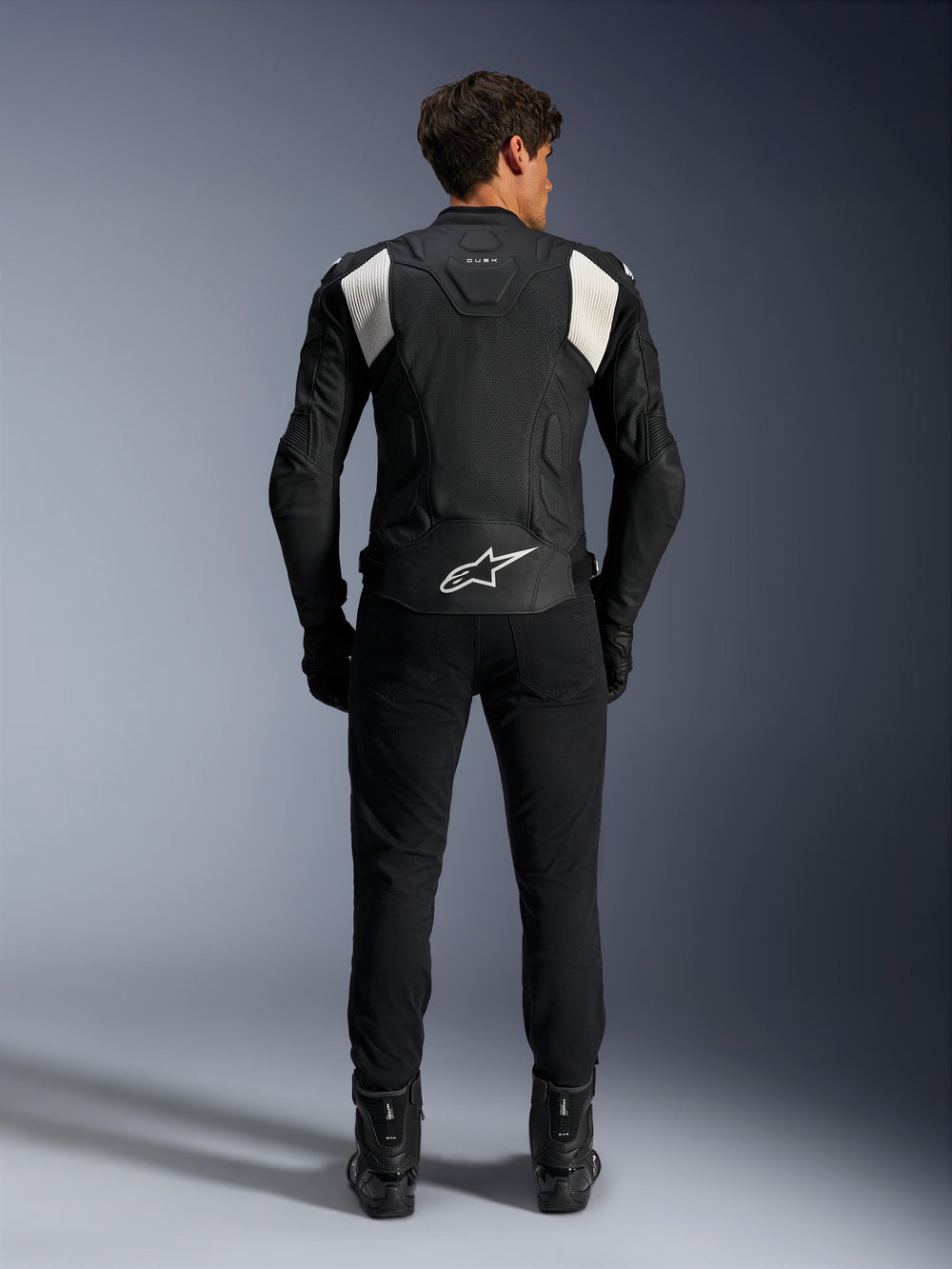 Alpinestars Dusk Airflow Leather Jacket, Black and White, rear view of a rider wearing black pants and boots, featuring perforated leather for ventilation, white accordion stretch panels on the shoulders, and white branding on the lower back