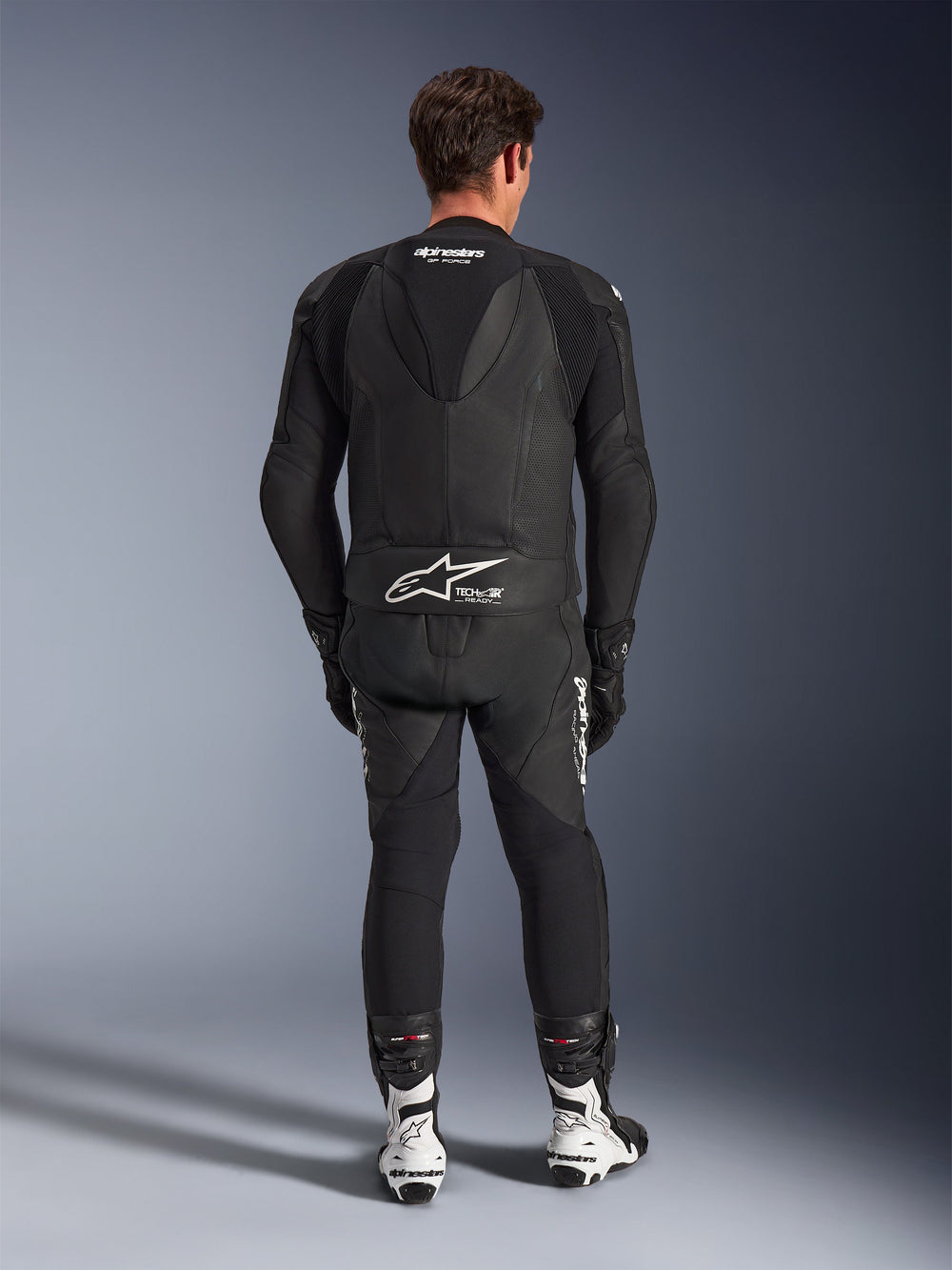 Alpinestars GP Force V2 Leather Suit 2-Piece, Black, back view showing aerodynamic speed hump and Tech-Air Ready branding on the waist, perforated leather panels for ventilation, paired with white and black racing boots, professional motorcycle racing gear
