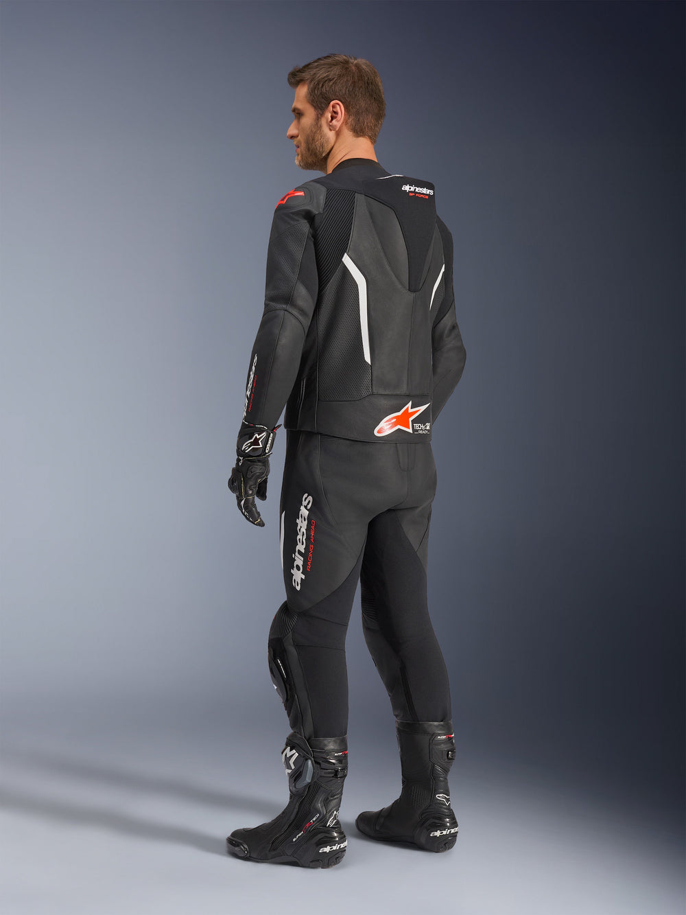 Alpinestars GP Force V2 2-Piece Leather Suit, Black with White and Red Fluo accents, rear three-quarter view on model, featuring an aerodynamic speed hump, perforated leather panels, and bold Alpinestars branding, worn with black racing boots and gloves.