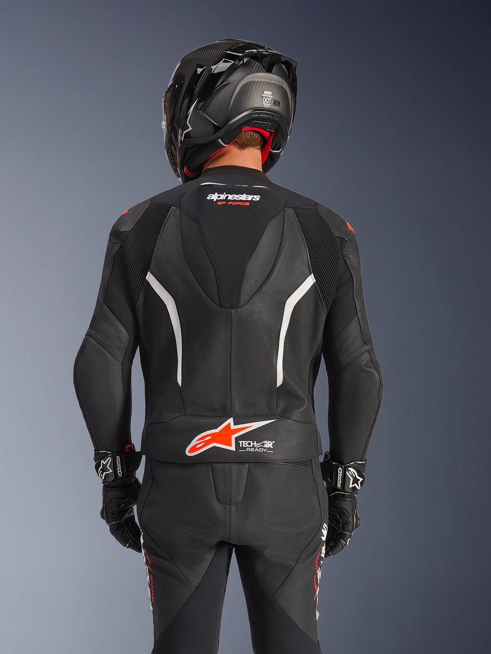 Rear view of Alpinestars GP Force V2 2-Piece Leather Suit in Black, featuring perforated leather, white accents, and red Alpinestars branding on the lower back, worn with a black motorcycle helmet and racing gloves.