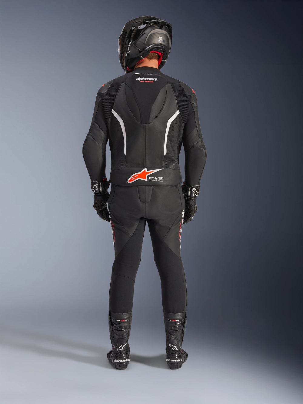 Motorcycle rider from behind wearing the Alpinestars GP Force V2 2-Piece Leather Suit, black with white and red fluo accents, featuring an aerodynamic hump and Tech-Air Ready logo, complete with a black racing helmet, gloves, and boots.