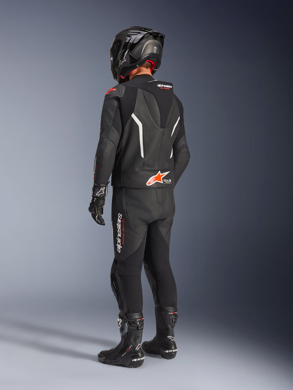 Alpinestars GP Force V2 Leather Suit - 2Piece, Black motorcycle leather suit seen from the back, worn with a matching black helmet, gloves, and boots, featuring white and red accents, aerodynamic speed hump with Alpinestars branding, professional racing and sport riding gear.