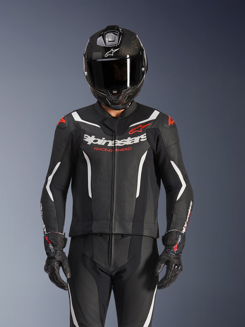 Alpinestars GP Force V2 2-Piece Leather Suit, Black White and Red Fluo, worn by a rider with a carbon fiber full-face helmet and black racing gloves, featuring perforated leather panels and Alpinestars branding on the chest.