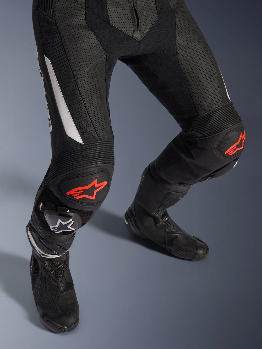 Alpinestars GP Force V2 2-Piece Leather Suit, motorcycle racing pants in black with red and white accents, featuring perforated leather, accordion stretch panels above the knees with red star logos, worn with black motorcycle boots, high-performance racing gear