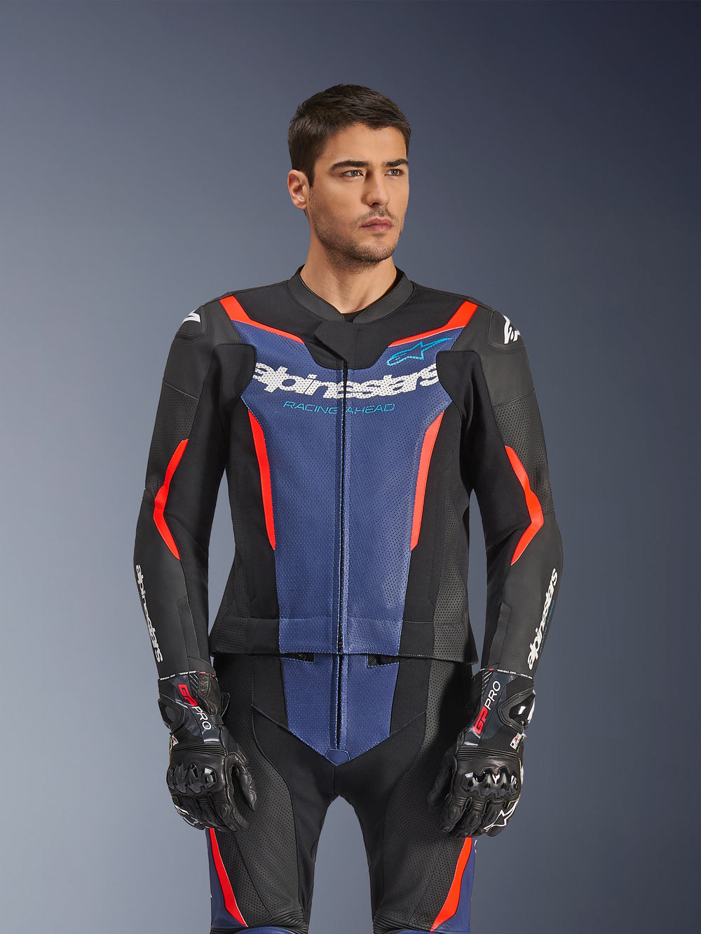 Male model wearing Alpinestars GP Force V2 Leather Suit 2-Piece, Black Blue Red Fluo, featuring bold blue chest panel and red accents, worn with GP Pro gloves, professional leather motorcycle racing gear with perforated texture