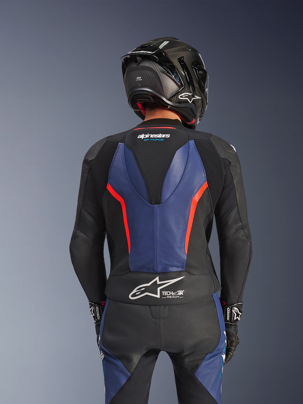 Alpinestars GP Force V2 2-Piece Leather Suit, Black Blue Red Fluo, rear view showing aerodynamic speed hump and Tech-Air Ready branding, premium bovine leather with stretch zones, worn with a black carbon fiber helmet, professional motorcycle racing gear