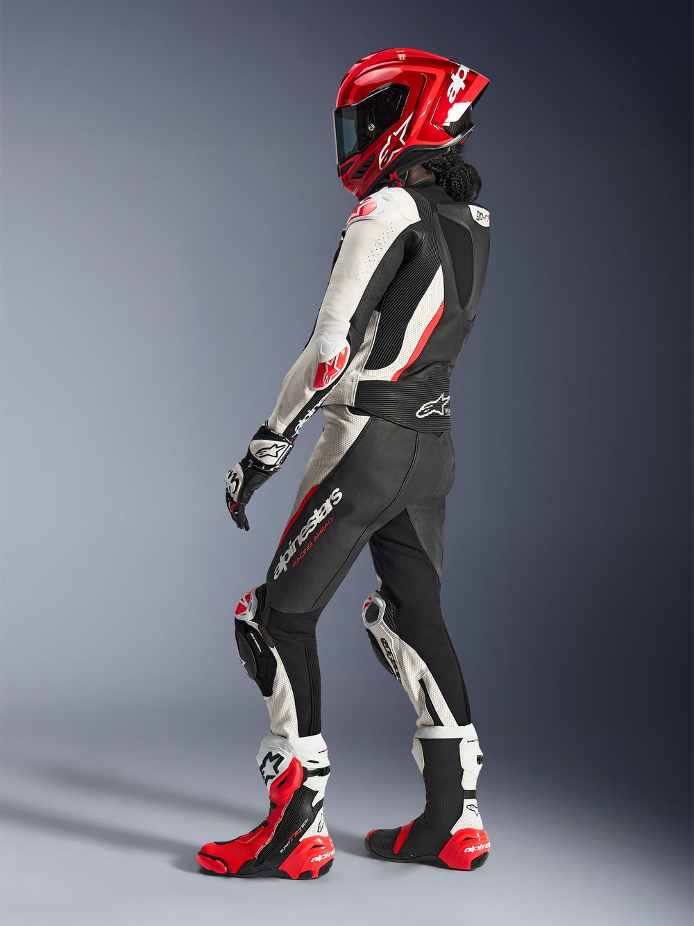 Woman rider wearing Alpinestars Stella GP-R7 2-Piece Leather Suit, Black White and Red Fluo, rear-three-quarter view showing aerodynamic hump, matching red full-face helmet, black and white racing gloves, and red and white technical racing boots, professional motorcycle racing gear