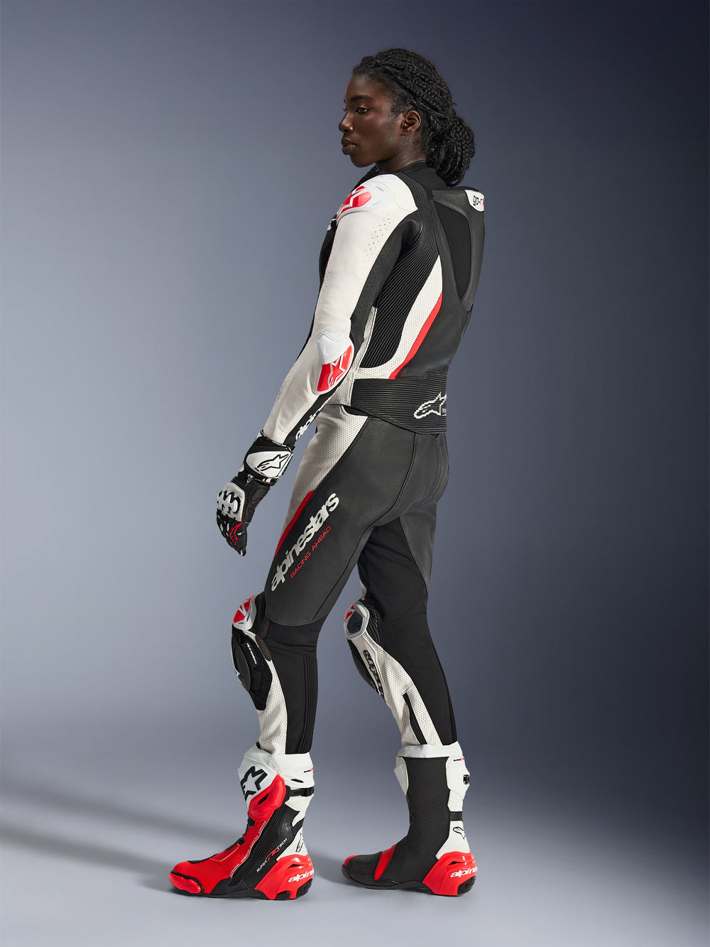 Woman wearing Alpinestars Stella GP-R7 2-Piece Leather Suit, Black White and Red Fluo, side profile showing aerodynamic back hump and knee sliders, paired with matching Alpinestars racing gloves and boots, professional motorcycle track gear