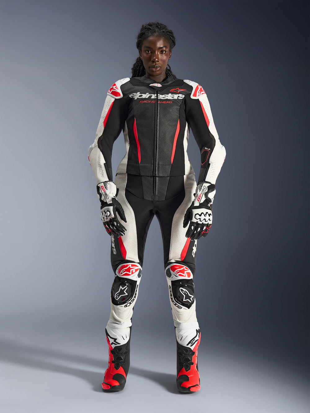 Woman model wearing Alpinestars Stella GP-R7 2-Piece Leather Suit in Black, White, and Red Fluo, complete with matching gloves and racing boots, featuring protective armor and perforated leather panels, professional motorcycle racing gear