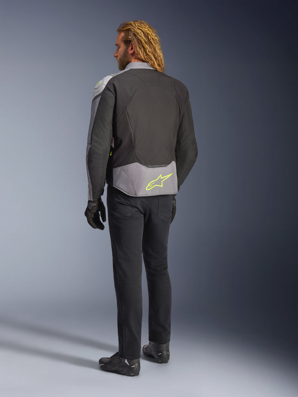Alpinestars T-Jaws V4 Waterproof Jacket, back view, Black Ice Gray and Dark Gray, featuring a neon yellow Alpinestars logo on the lower back, worn with black pants and motorcycle gloves, sport-styled riding apparel
