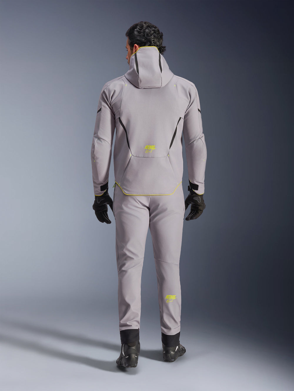 Alpinestars Aeroshell Waterproof Jacket in Mid Gray Yellow Fluo, full back view, worn with matching gray pants, black gloves, and black riding shoes. Features a hooded softshell design with yellow fluo 'ASTARS' branding and accents, engineered for modern motorcycle road performance.