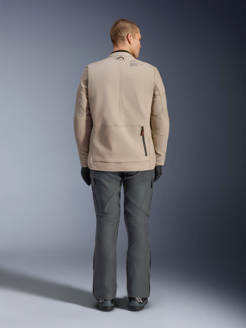 Alpinestars Borrego Drystar® Jacket, back view of male model wearing the Warm Gray textile motorcycle jacket, paired with dark gray technical pants, black gloves, and boots, featuring the rugged waxed-canvas design and rear expansion panels.