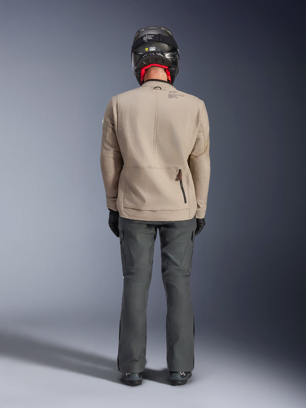 Alpinestars Borrego Drystar® Jacket, Warm Gray adventure motorcycle jacket, rear view showing waxed-canvas look with shoulder expansion panels and lower back zipper pocket, worn with dark gray riding pants and black helmet.