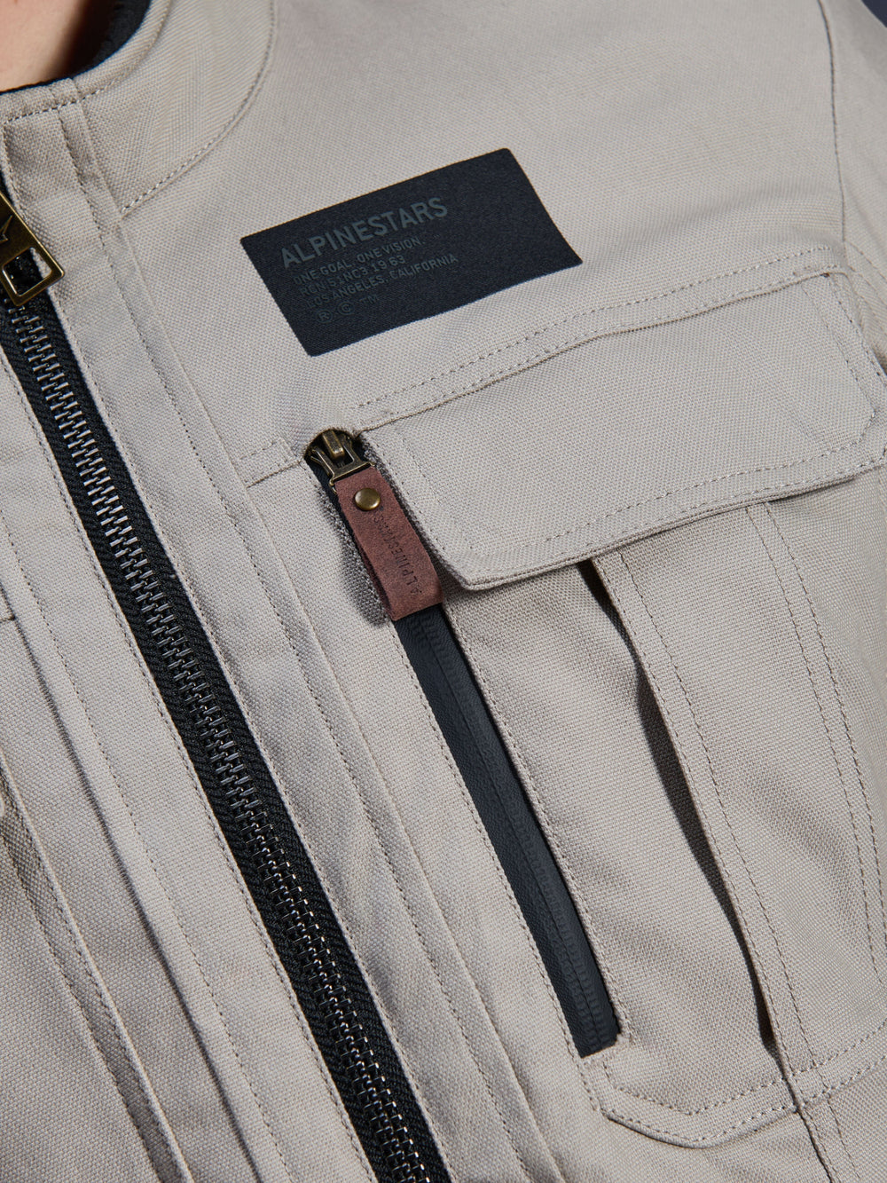 Alpinestars Borrego Drystar® Jacket, Warm Gray, close-up detail of chest area featuring a black Alpinestars branding patch, chest pocket with flap, and a vertical zipper with a brown leather pull tab, durable gray textile canvas construction.