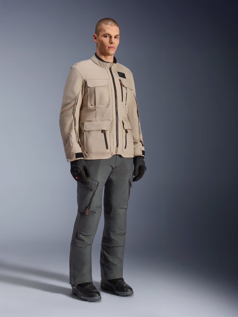 Alpinestars Borrego Drystar® Jacket in Warm Gray, adventure motorcycle jacket with a waxed-canvas look and multiple utility pockets, worn by a model with dark gray riding pants, black gloves, and black boots, full-body studio portrait
