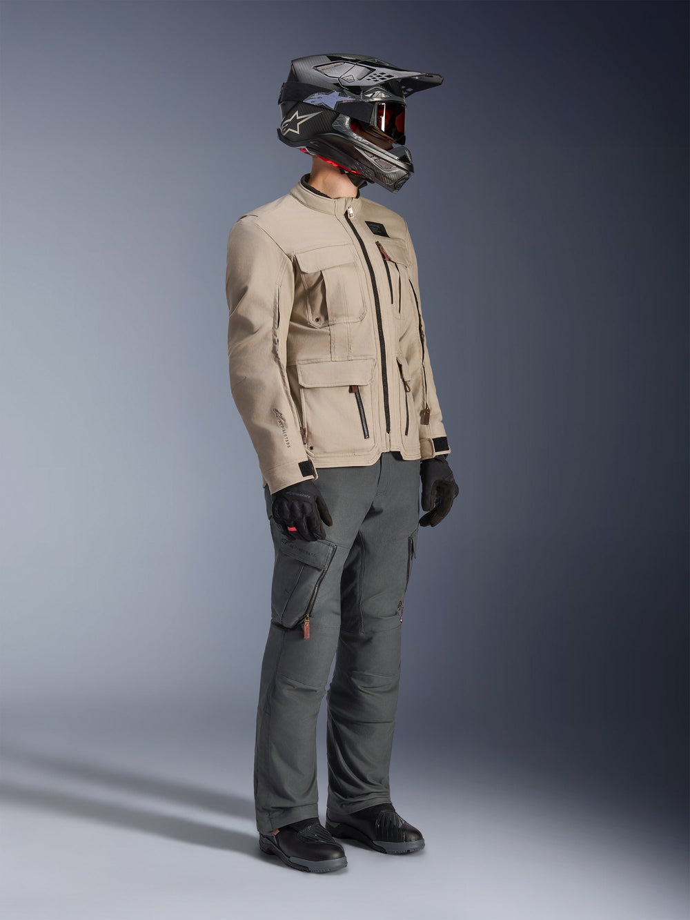 Motorcyclist wearing Alpinestars Borrego Drystar® Jacket in Warm Gray, adventure motorcycle jacket with a waxed-canvas texture and utility pockets, styled with dark gray riding pants, a carbon fiber helmet, black gloves, and black boots, full-body studio view of a complete adventure touring outfit.
