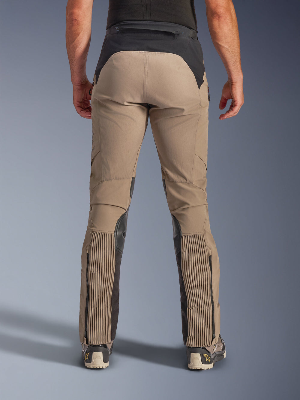 Alpinestars AMT-8 Stretch Drystar® XF Pants, Walnut Black, rear view on model, tan stretch textile chassis with black waist yoke and calf panels, featuring large accordion stretch sections on the lower legs, zippered leg entries, and adventure-touring fit