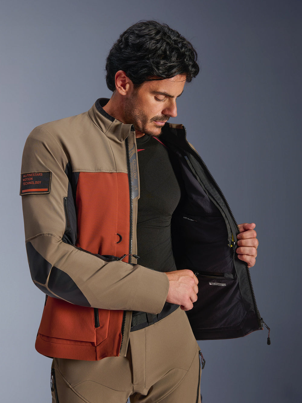 Alpinestars AMT-8 Stretch Drystar® XF Jacket, Walnut Rust Black, man wearing the jacket open to show the interior waterproof compartment and technical lining, featuring 'Alpinestars Motion Technology' patch on the arm, adventure motorcycle gear.