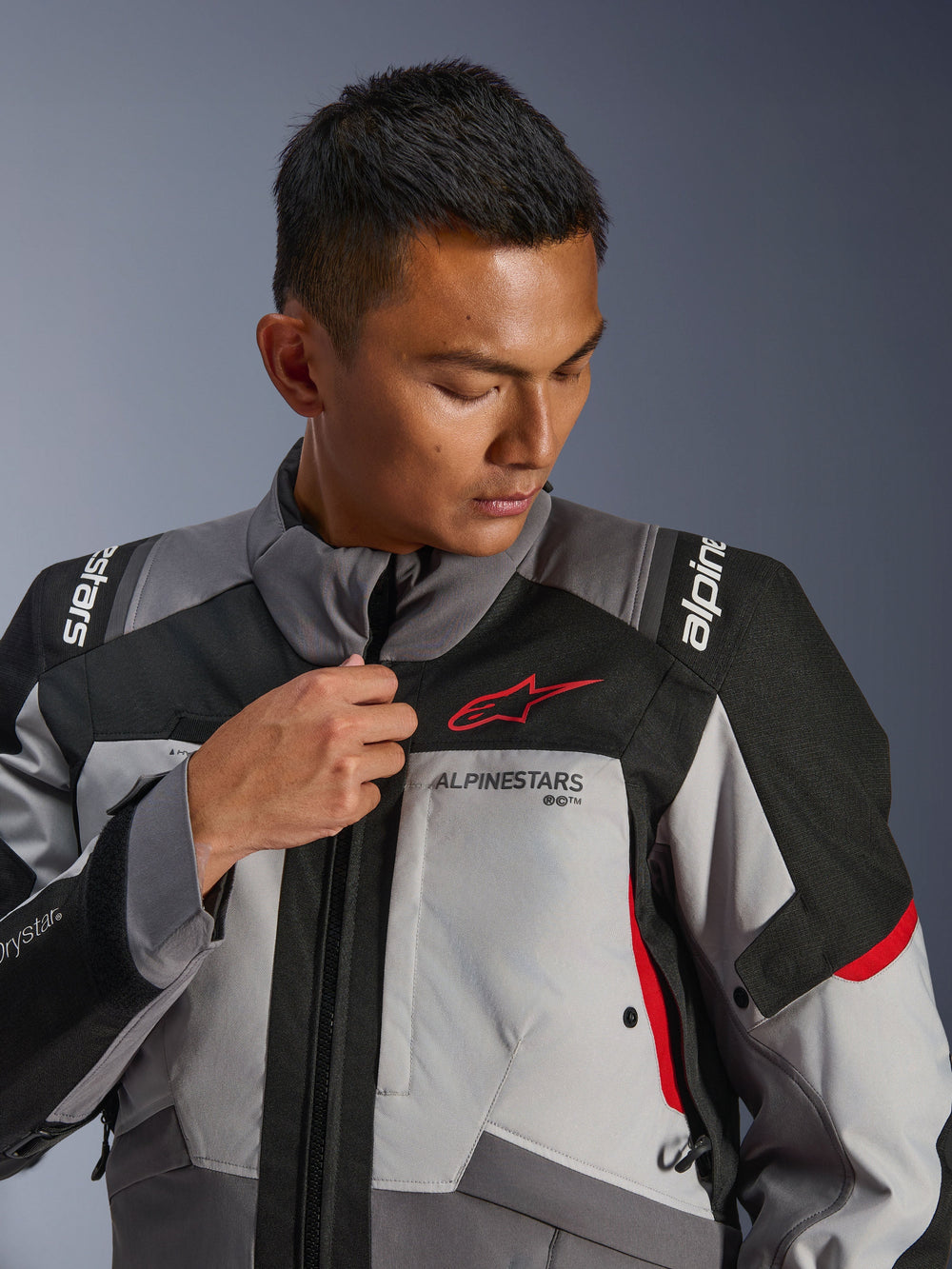 Model wearing Alpinestars Andes V4 Drystar® Jacket, Dark Gray and Ice Gray with Black and Bright Red accents, adjusting the front zipper, featuring the red Astars logo and Drystar® branding, adventure touring style