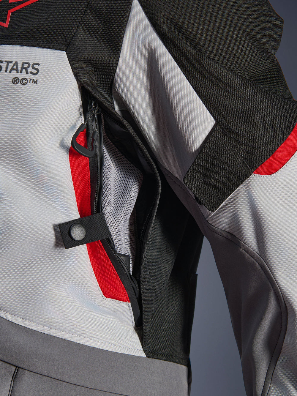 Close-up detail of the Alpinestars Andes V4 Drystar® Jacket in Dark Gray, Ice Gray, and Bright Red, highlighting a zippered chest ventilation port with mesh lining and a snap button adjustment strap.