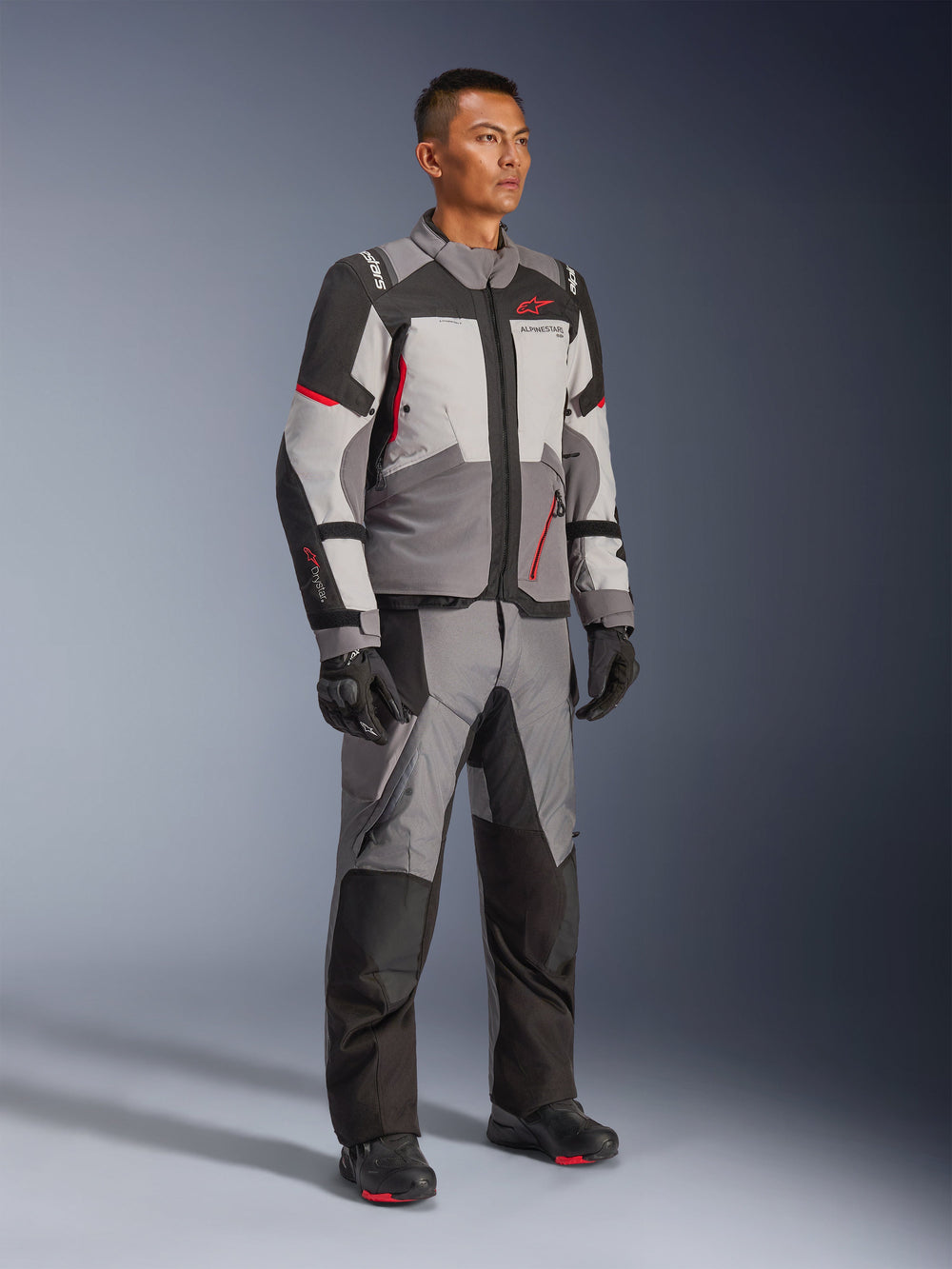 Male model wearing Alpinestars Andes V4 Drystar® Jacket and matching adventure pants, Dark Gray, Ice Gray, Black, and Bright Red, paired with black motorcycle gloves and boots, technical all-weather riding gear with a modern multi-panel design.