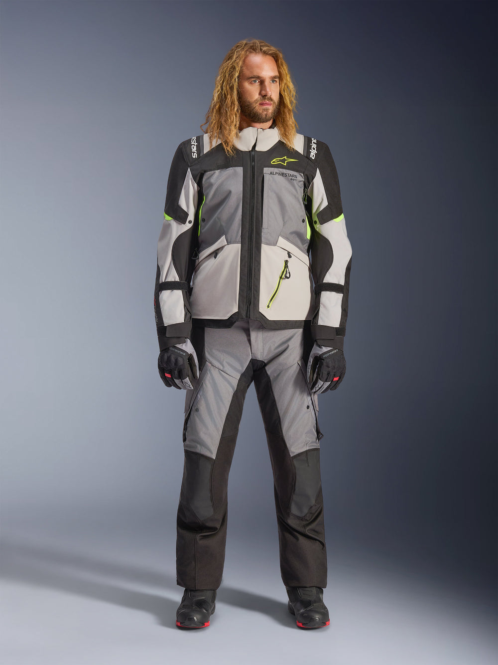 Male model with long blonde hair wearing the Andes V4 Drystar® Jacket in Dark Gray, Ice Gray, Black, and Yellow Fluo, paired with matching adventure motorcycle pants and black boots, full technical riding outfit for all-weather touring.