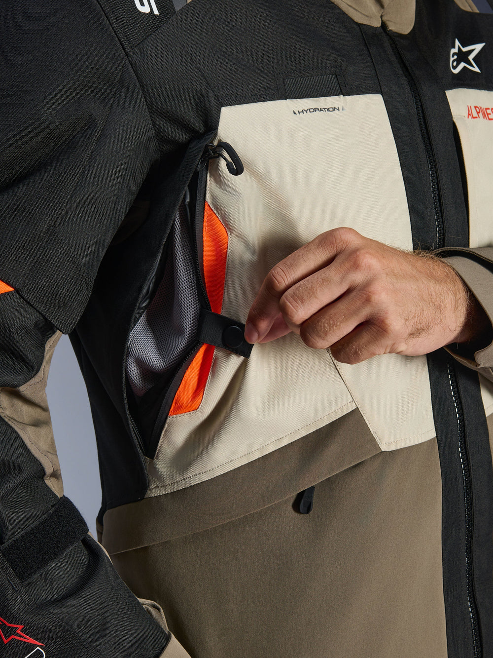 Alpinestars Andes V4 Drystar® Jacket detail shot, Walnut Mastic Black textile, close-up of hand opening the chest ventilation panel to reveal a bright orange mesh interior, showcasing the direct airflow system for adventure riding