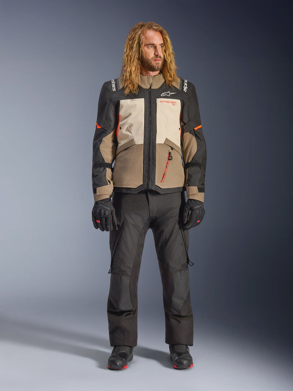 Alpinestars Andes V4 Drystar® Jacket, Walnut Mastic Black, worn with black motorcycle pants and gloves, featuring a multi-panel adventure design with orange accents and Alpinestars branding, technical textile gear for all-weather riding