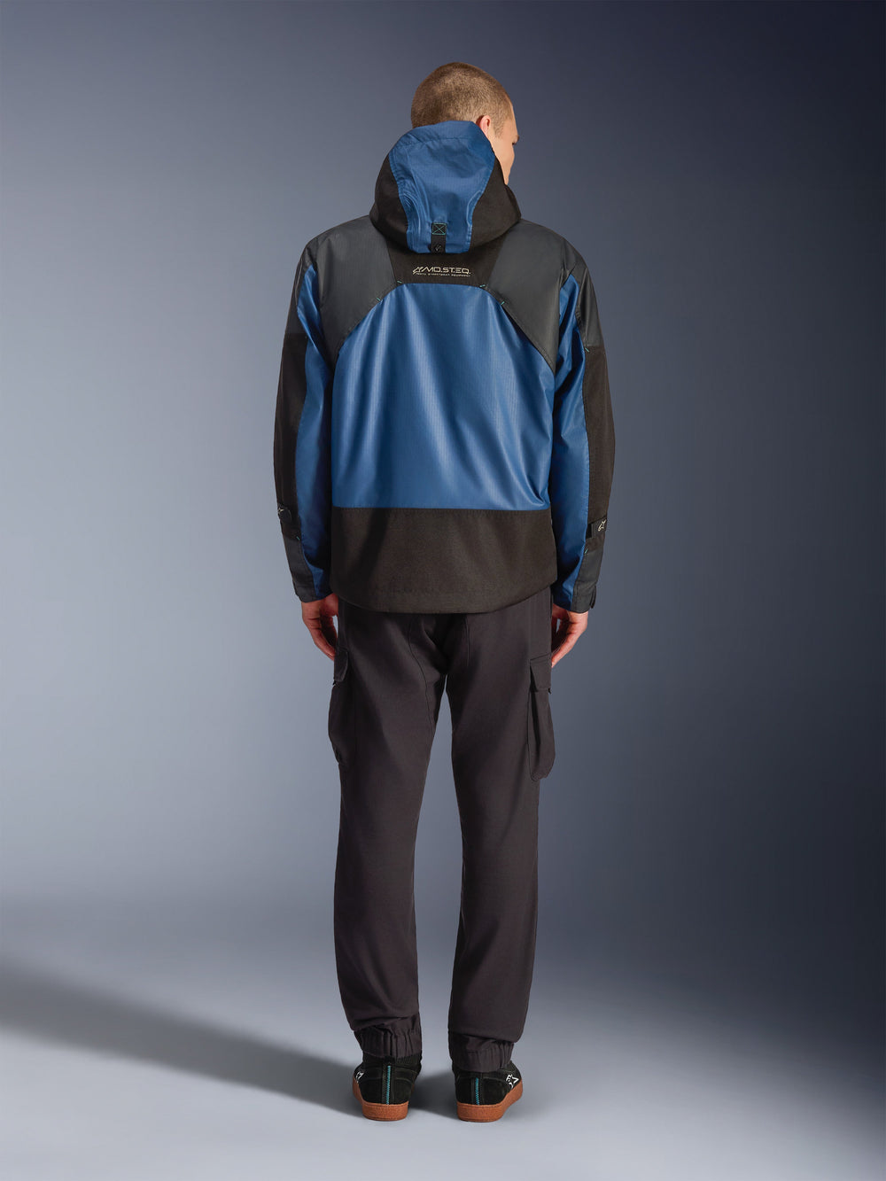 Alpinestars Mospher Waterproof Jacket in Black and Dark Petrol, rear view on model, paired with black cargo pants, featuring tactical ripstop construction and MO.ST.EQ. branding on the back.