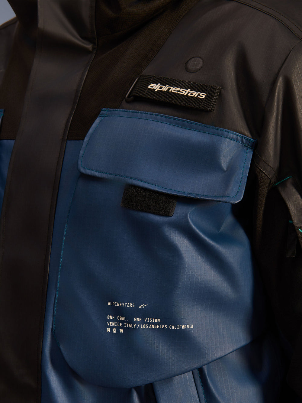 Alpinestars Mospher Waterproof Jacket, close-up detail of the chest pocket area, Black and Dark Petrol, durable ripstop fabric with printed 'One Goal. One Vision.' branding, utility-driven design with functional pockets and Alpinestars logo tab