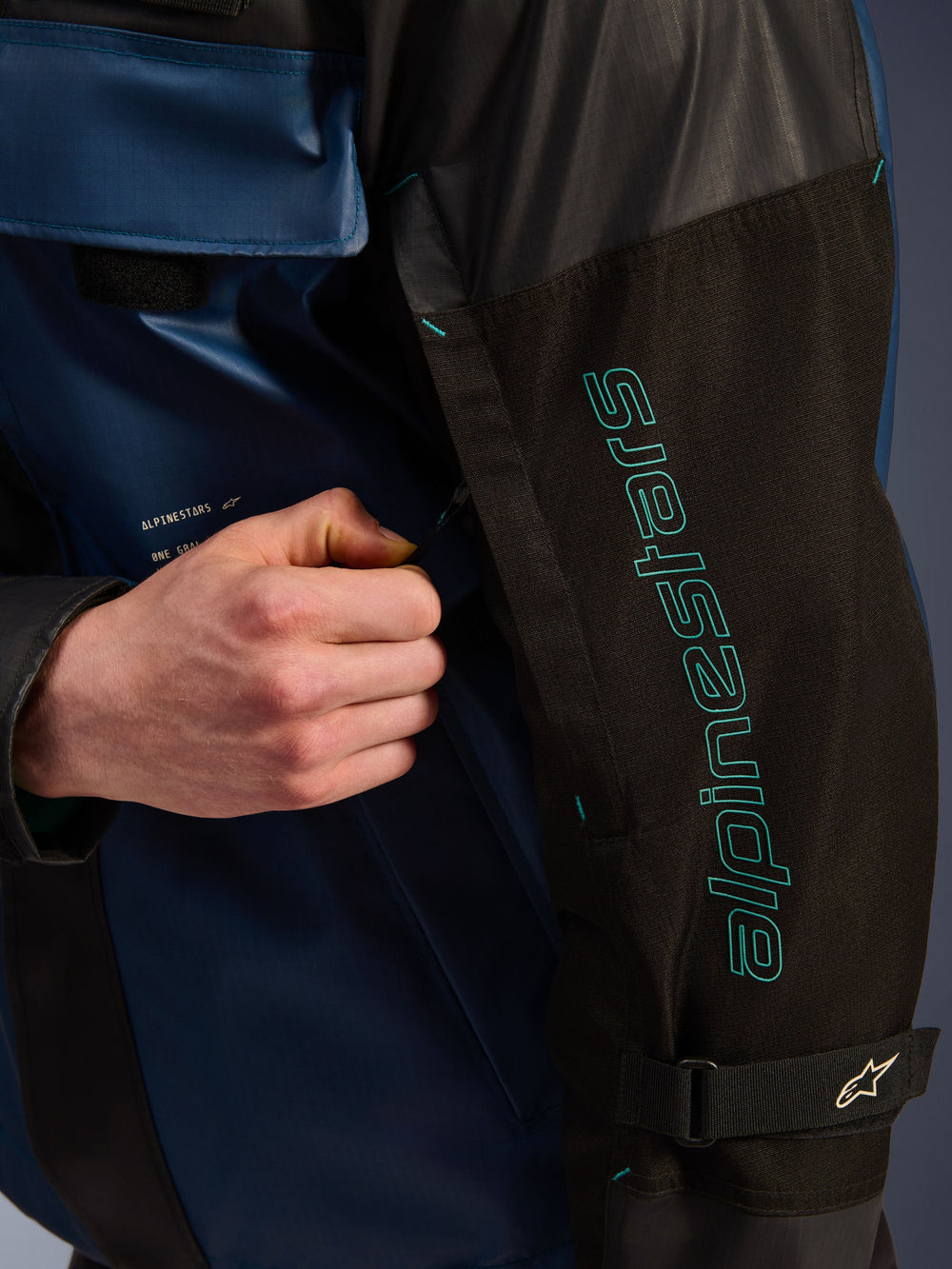 Alpinestars Mospher Waterproof Jacket in Black Dark Petrol, close-up detail of the sleeve with teal Alpinestars branding, a hand adjusting the ventilation zipper, featuring durable ripstop fabric and an adjustable arm strap