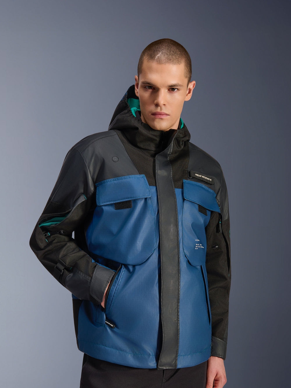 Male model wearing the Alpinestars Mospher Waterproof Jacket in Black and Dark Petrol, featuring large utility chest pockets, a hooded design with teal lining, and technical ripstop fabric for urban motorcycle commuting.
