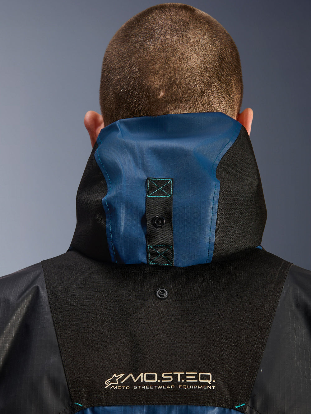 Alpinestars Mospher Waterproof Jacket, back view detail showing the hood anchoring system, Black and Dark Petrol Blue, featuring MO.ST.EQ. Moto Streetwear Equipment branding, durable ripstop textile material, technical commuter motorcycle gear