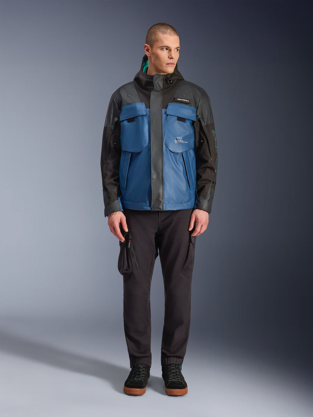 Alpinestars Mospher Waterproof Jacket in Black Dark Petrol, male model wearing tactical-inspired all-season motorcycle parka with large functional pockets and integrated hood, paired with black technical pants and black footwear, studio front view