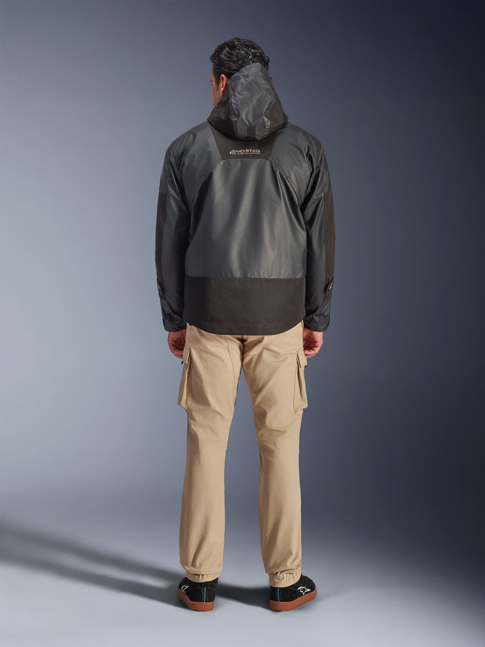 Alpinestars Mospher Waterproof Jacket, Black motorcycle parka shown from the back, featuring MO.ST.EQ logo on upper back and ripstop fabric construction, worn with tan cargo pants and black sneakers, urban commuter style.