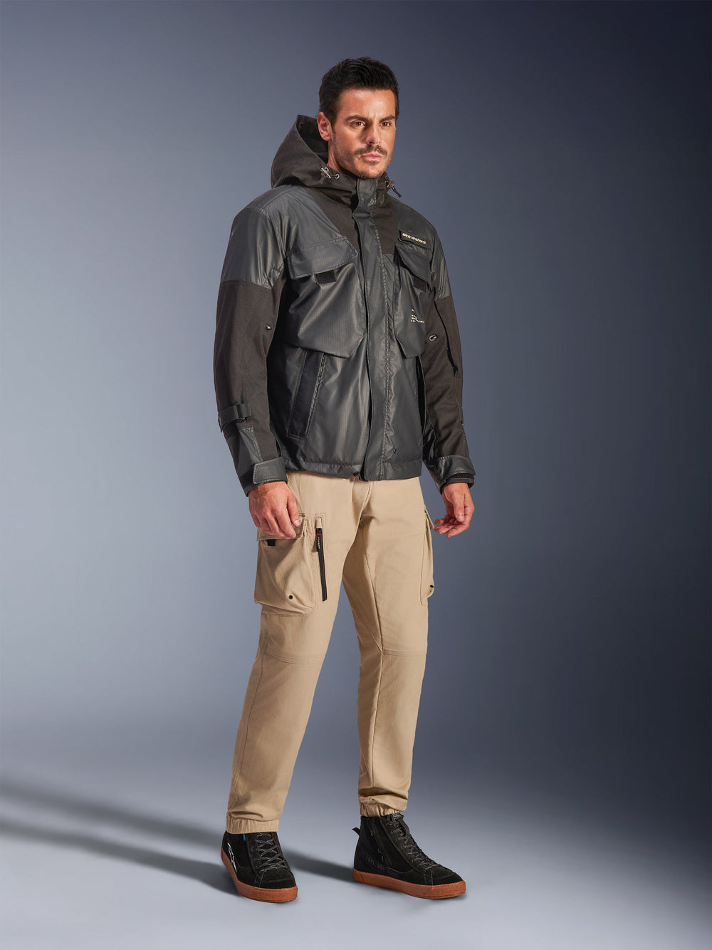 Male model wearing the Alpinestars Mospher Waterproof Jacket in Black, utility-inspired waterproof parka with a multi-pocket design and hood, paired with tan cargo pants and black high-top sneakers, urban motorcycle commuter gear.